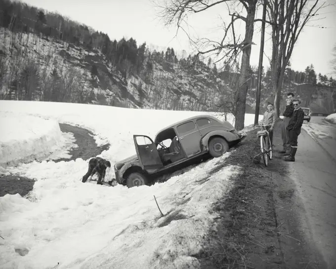 Vintage photograph. Man digging out car in snow filled ditch with onlookers