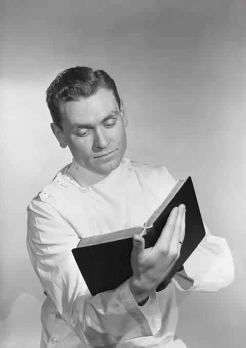 Vintage photograph. Man in white medical smock holding up book while reading