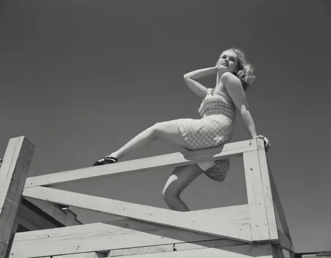 Woman in short dress sitting on railing of deck looking over shoulder
