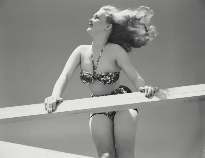 Woman in bikini leaning over railing of deck with hair blowing in wind