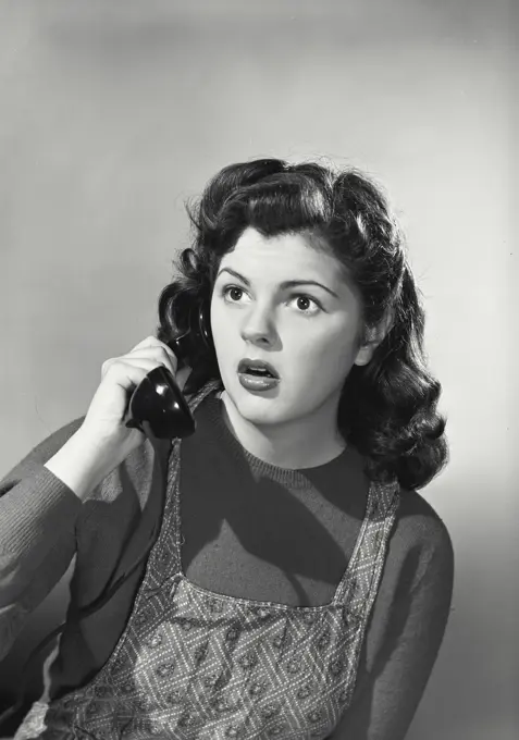 Vintage photograph. Portrait of young woman in apron talking on telephone with shocked expression on face