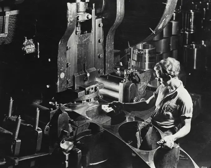 Vintage photograph. Man feeding aluminum sheets into stamping machine for cutting into circular pieces for working