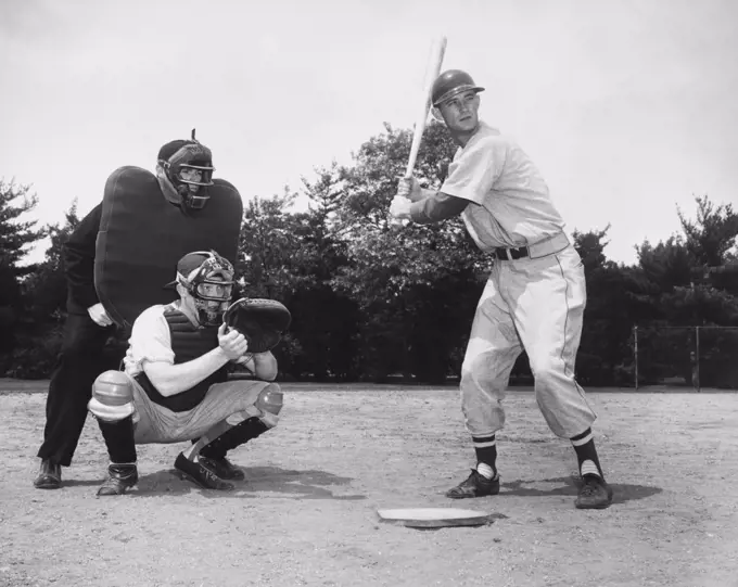 Baseball player swinging a baseball bat with a baseball catcher and a baseball umpire crouching beside him