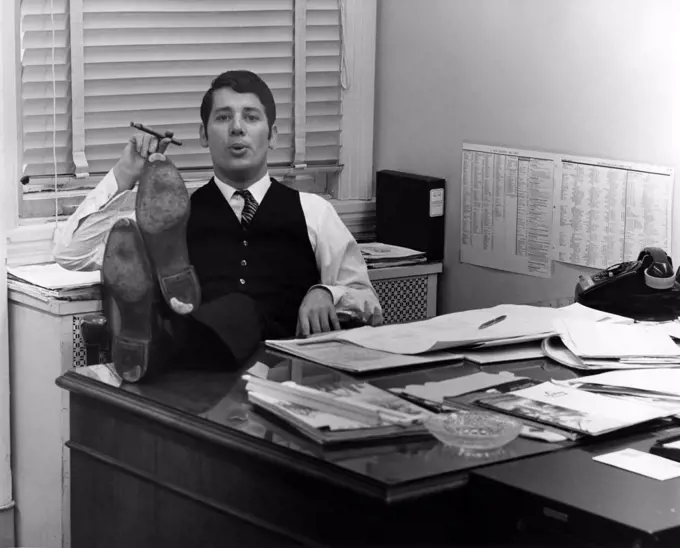 Clerk sitting in office room with legs on table, smoking cigar