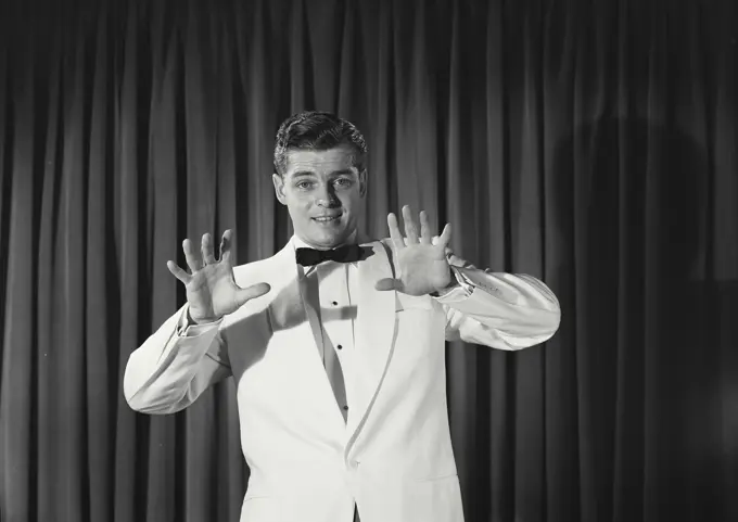 Vintage Photograph. Young man wearing white tuxedo holding up hands in front of wall of curtains