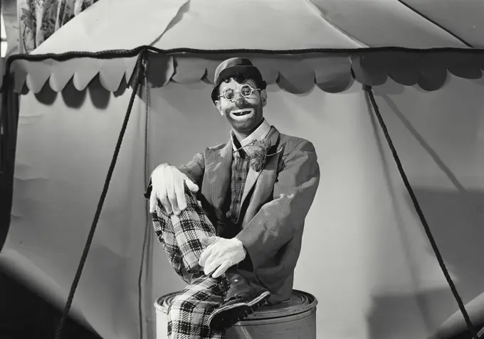 Vintage Photograph. Hobo clown crossing legs sitting on barrel in front of circus tent