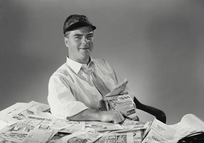 Vintage Photograph. Businessman wearing visor sitting back at office desk covered in newspaper clippings smiling at camera