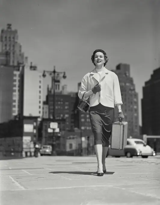 Vintage Photograph. Nicely dressed woman walking down sidewalk carrying purse and small suitcase