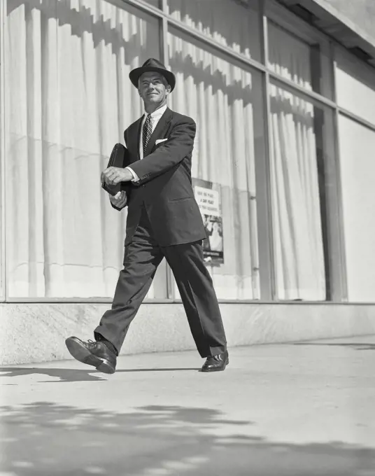Bill Hammond Model Released. Vintage Photograph. Older man wearing suit and hat carrying thin leather briefcase walking outside on sidewalk by building with large windows
