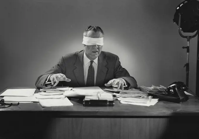 Vintage Photograph. Blindfolded man sitting at desk covered in documents. Frame 1
