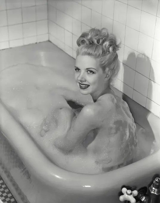 Vintage Photograph. Smiling blonde woman sitting up in bathtub covered in soap suds