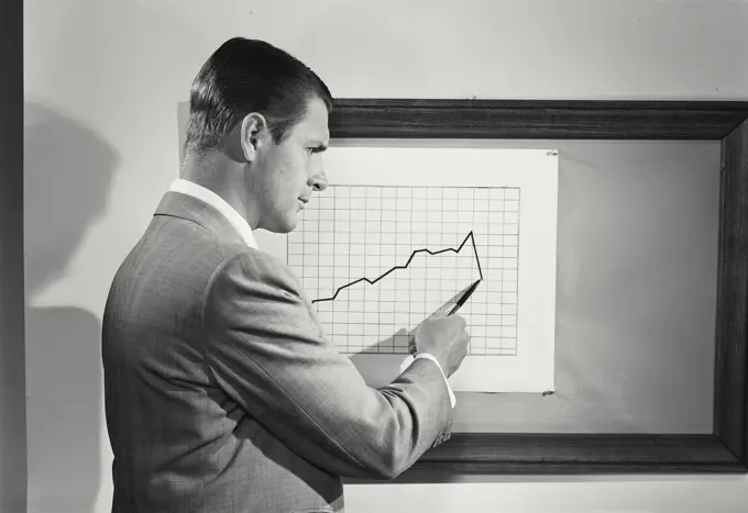 Vintage Photograph. Man points to chart showing declining numers as he looks unhappily to right of frame.