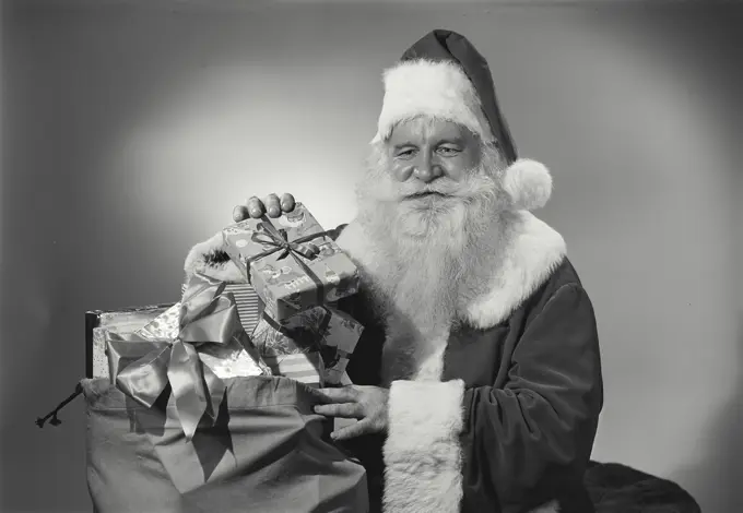 Vintage Photograph. Man in Santa Claus outfit takes gifts out of sack as he smiles.