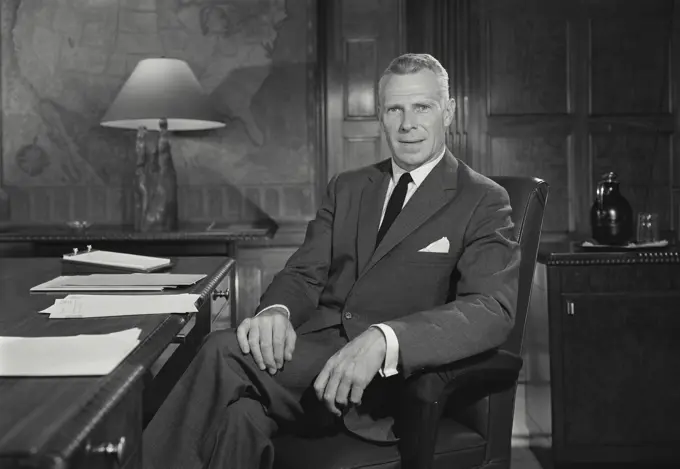 Vintage Photograph. Man sits at desk facing camera with legs crossed.