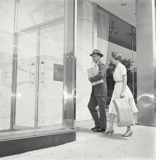 Vintage Photograph. Man and woman walk into front of bank building man carries packages while woman carries a bag.