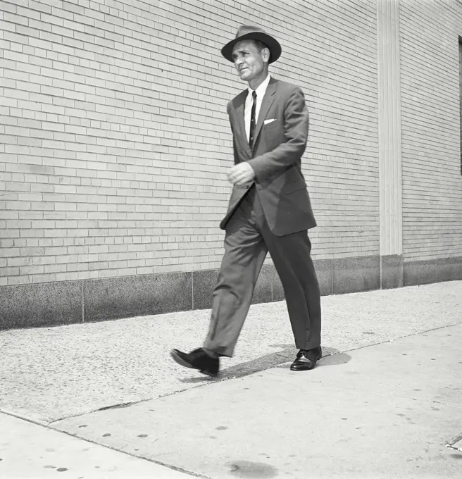 Vintage Photograph. Man walks down city sidewalk wearing suit tie and hat.