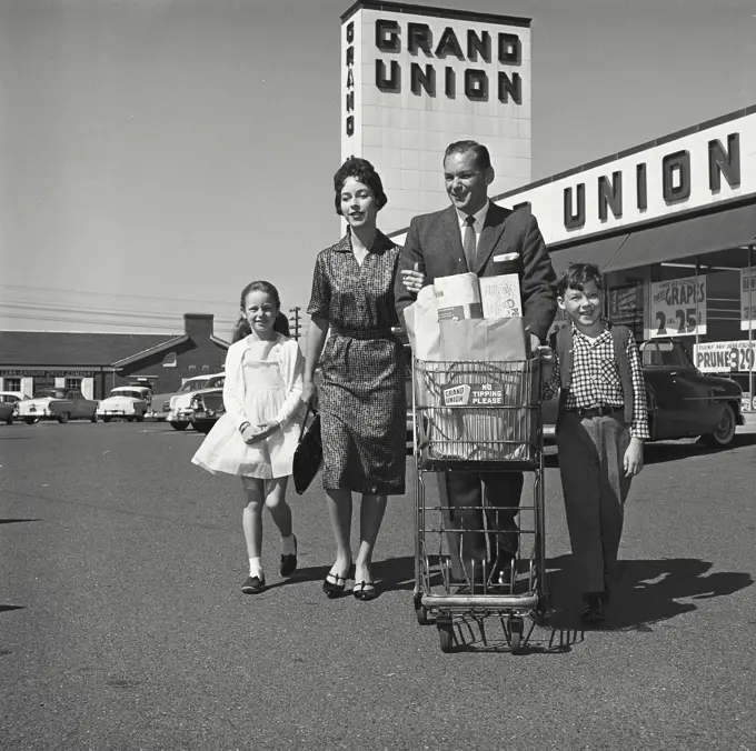 Vintage Photograph. Man pushes family's grocery cart in parking lot as the family leaves the grocery store.