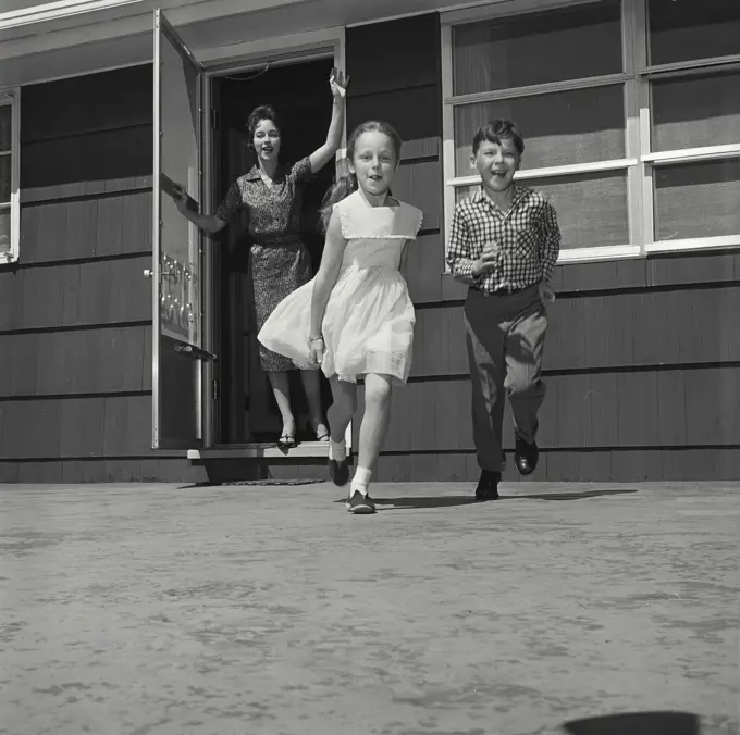 Vintage Photograph. Woman waves as two kids run towards the camera from front of house.