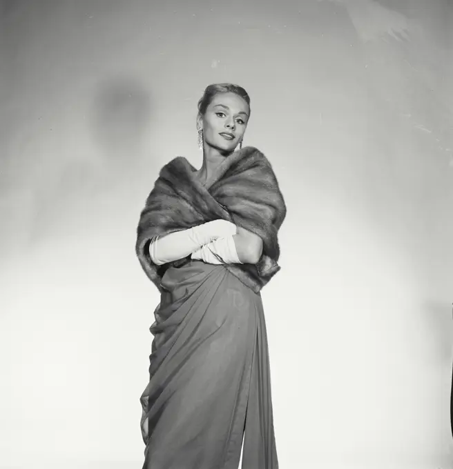 Vintage Photograph. Woman wears formal attire including dress and fur coat as she poses for studio portrait.