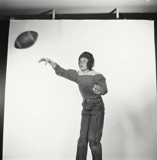 Vintage Photograph. Young boy throws football to the left of frame as he wears jeans and football uniform.