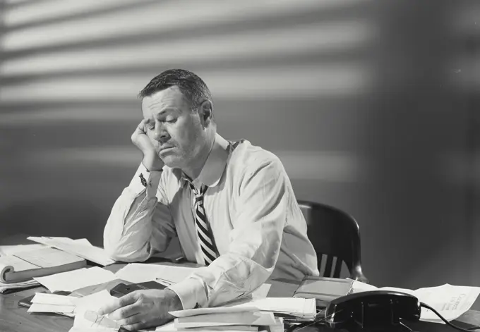 Vintage Photograph. Man in dress shirt and striped tie sitting at desk covered with papers looking exhausted