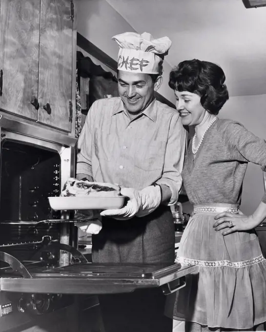 Mid adult man taking food out of an oven with a mid adult woman standing beside him