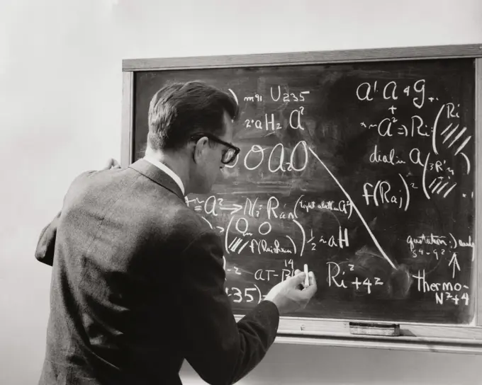 Rear view of a male teacher writing with chalk on a blackboard