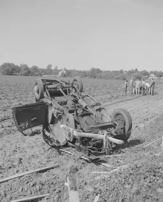 Overturned car in a field, crowd gathered near injured.