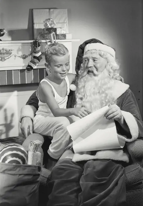 Vintage photograph. Young boy sitting with Santa Claus on chair in front of fireplace