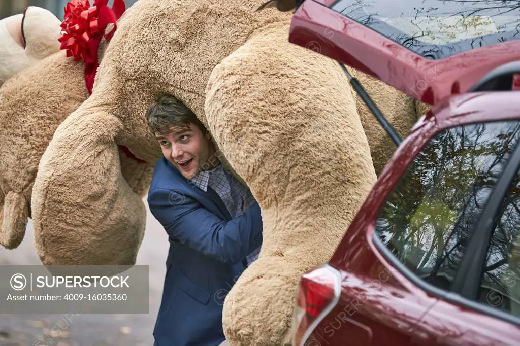 3/4 MCU of a young man grabbing a large teddy bear from the trunk of his car, throwing it over his shoulder and walking off camera.