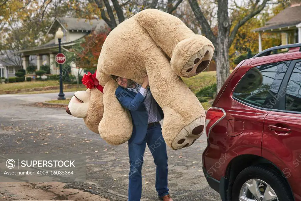 Wide shot of a man carrying a large teddy bear over his shoulder.