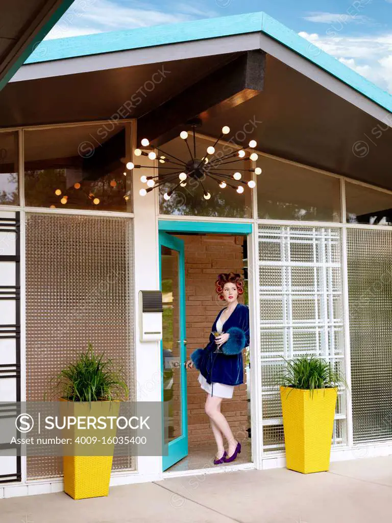 Vintage-styled woman standing outside the door of her mid-century home smiling.