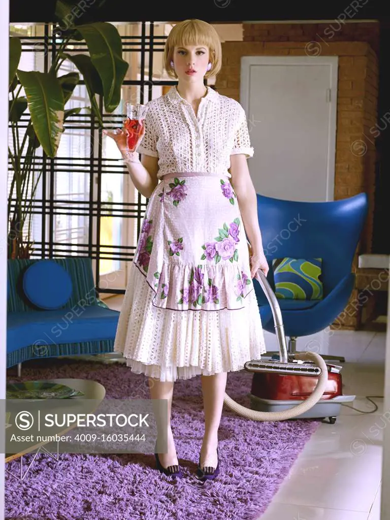 Woman dressed in vintage attire holding a vacuum in one hand and drink in the other in a mid-century modern home.