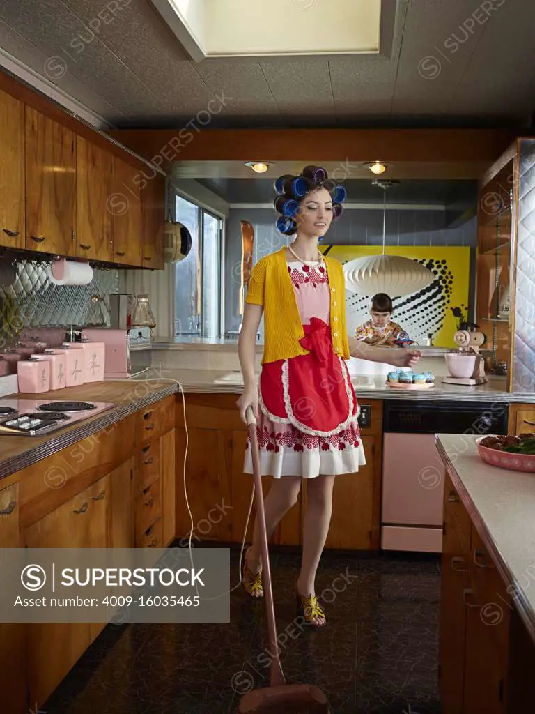 Woman vacuuming kitchen while wearing hair rollers in a mid-century style kitchen.