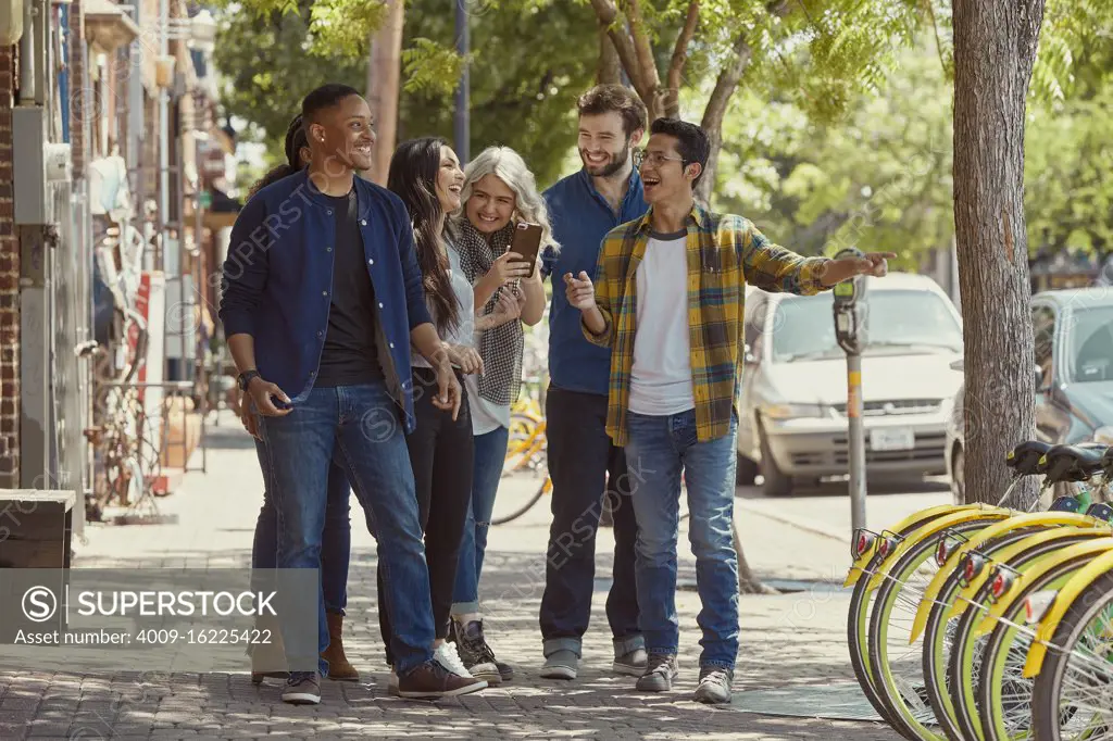 Group of young co-workers walking down sidewalk together, young woman showing friends something funny on cell phone 