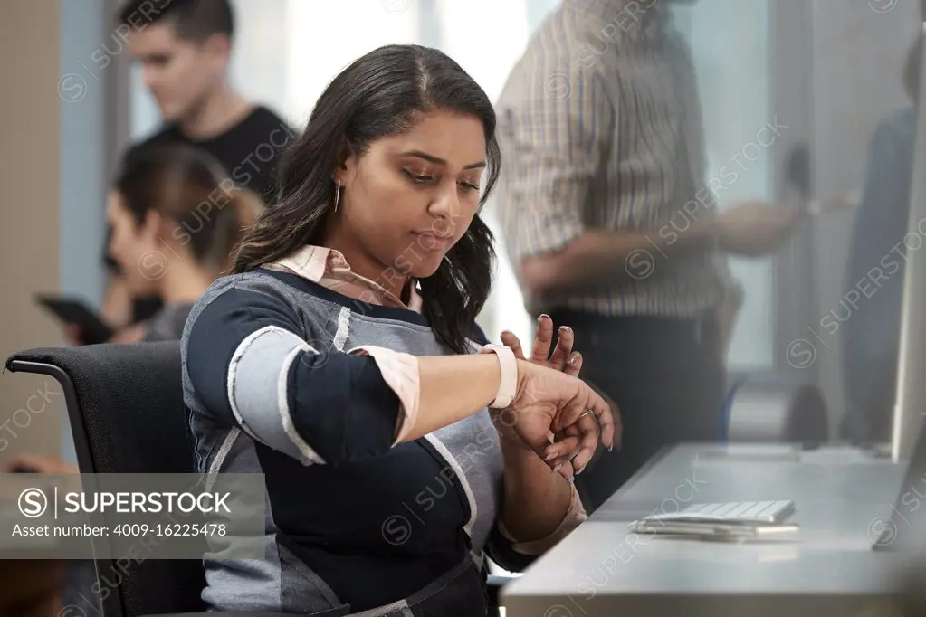 Young woman sitting at computer in office looking at smart watch, co-workers having discussion around dry erase board in background 