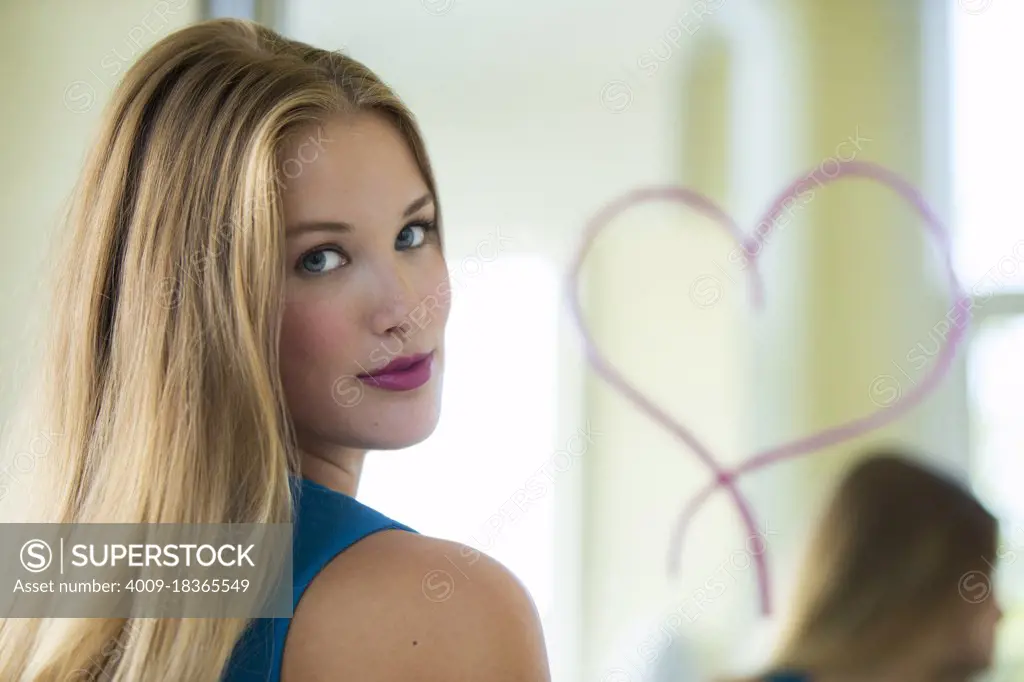 Portrait of young woman in bathroom, looking over shoulder to camera. In the background there is a mirror with a lipstick heart drawn on it. 