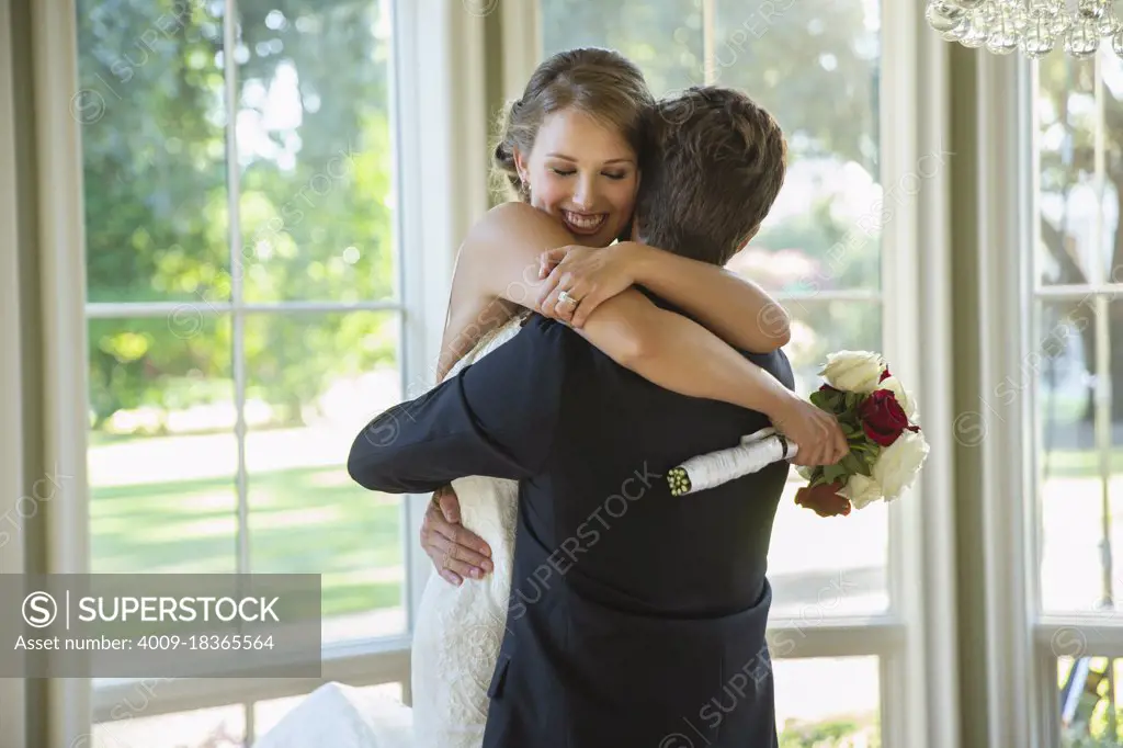 Bride and groom in an embrace, groom lifting up bride standing by large bank of windows 