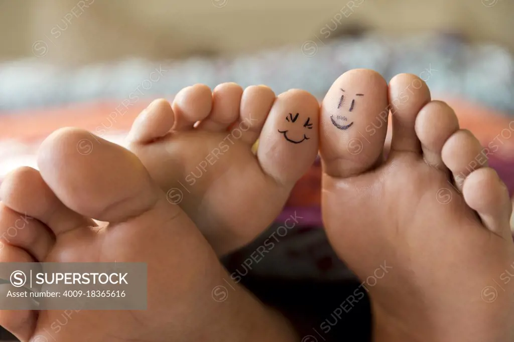 Close-up of bare feet sticking out from covers while couple lays in bed, with smiley faces drawn on big toes