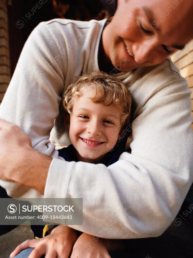 Father with arms wrapped around smiling little boy sitting on front steps of home