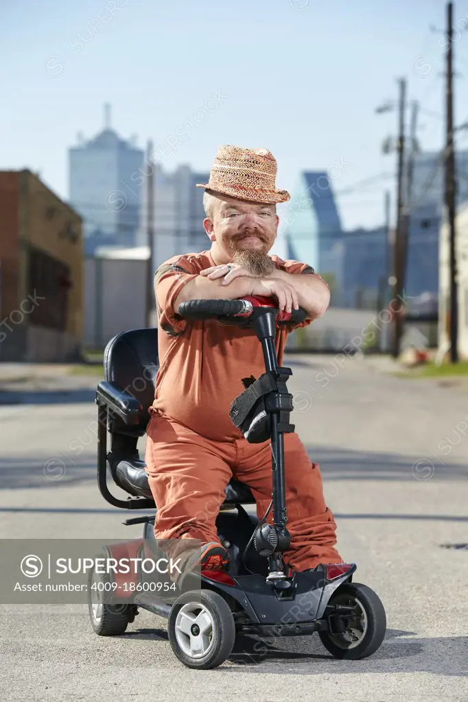 Portrait of male dwarf in sitting on scooter, looking into camera with big smile. 