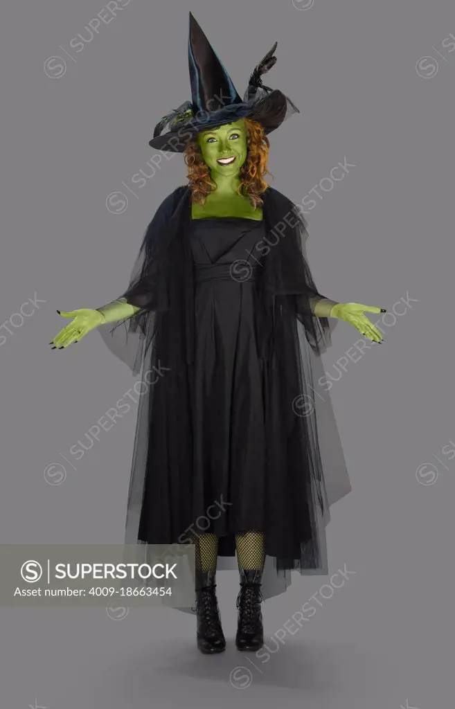 Full length portrait on gray of a woman with green body paint dressed up in a witch costume for Halloween with her arms out.