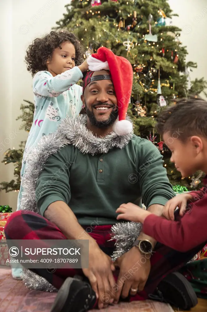 Man sitting in front of Christmas tree being decorated by his children