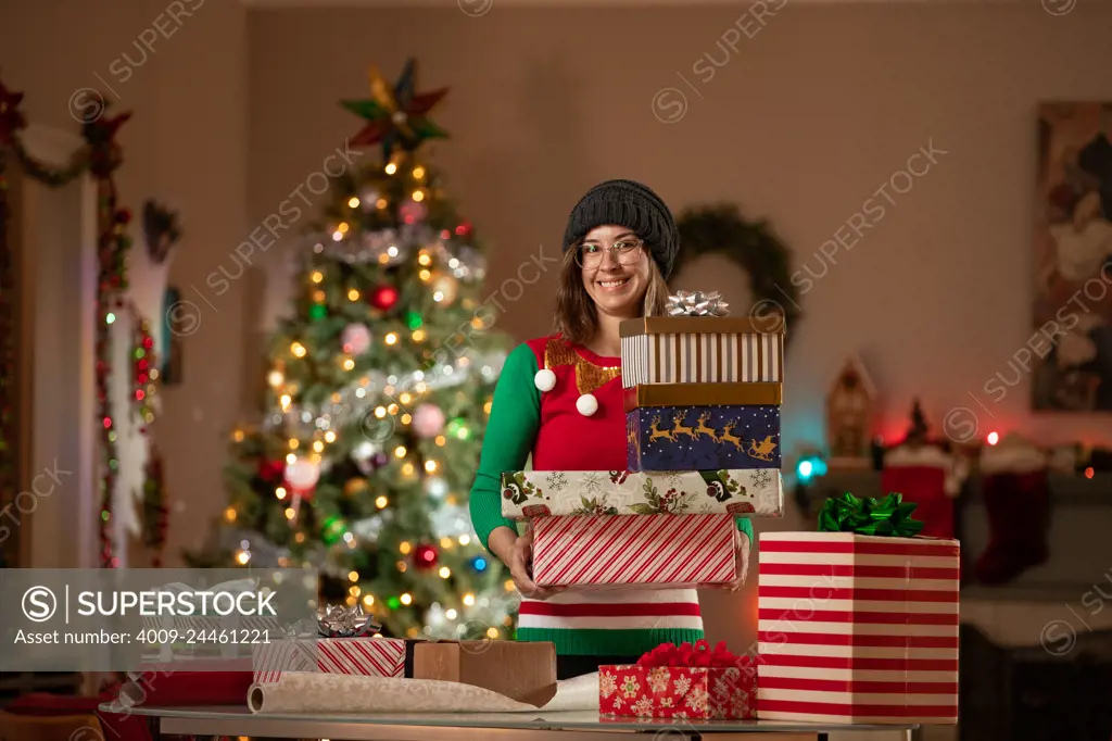 Young woman holding Christmas gifts in front of a Christmas Tree in her living room. 