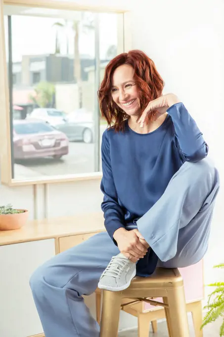 Medium shot of a middle-aged European looking woman sitting on a stool with a leg up, in a brightly lit room