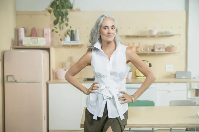 Portrait of woman over 40 standing in kitchen with hand on hips looking into camera.