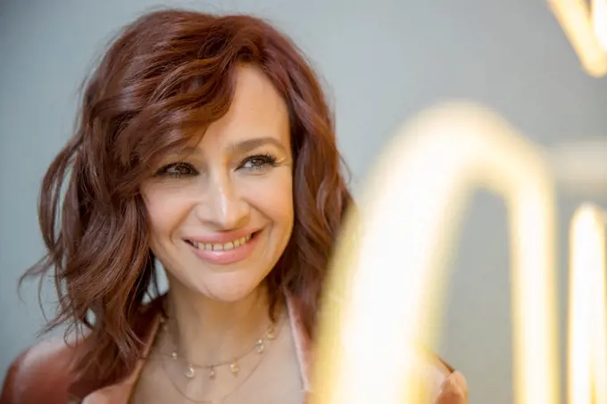 Casual portrait of woman with red hair standing next to neon sign looking off camera smiling