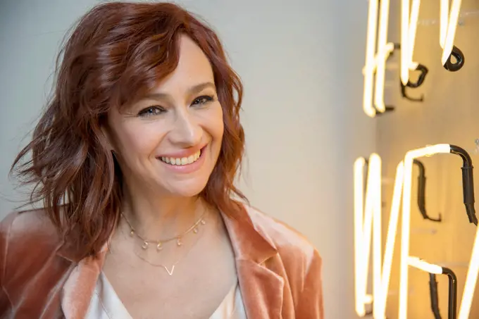 Middle-aged woman with red hair standing next to neon sign, looking into camera smiling.