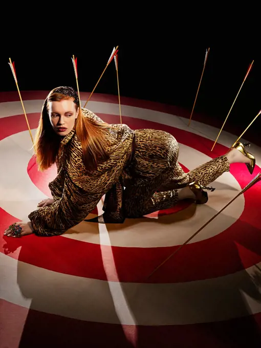 Portrait of young woman on her hands and knees crawling on a large hand painted Bulls-eye target with arrows and high heels