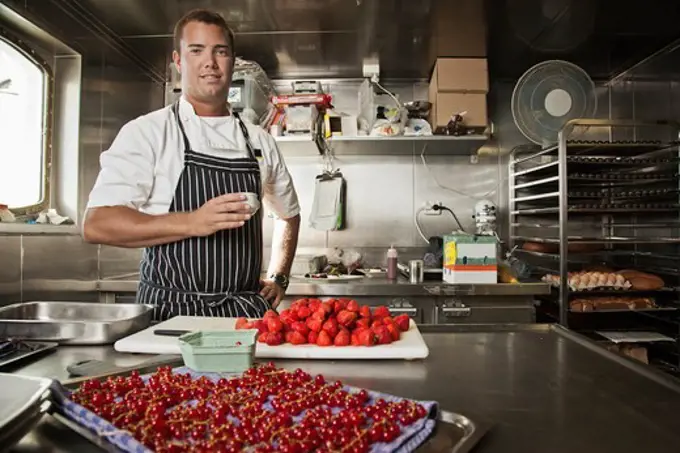 Pastry chef holding a cup of espresso while standing in kitchen with assorted berries being prepped on counter